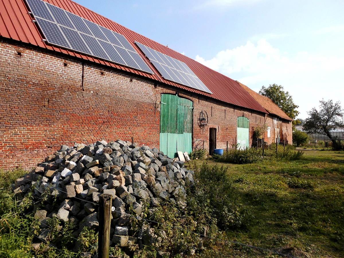 Oude schuur in rode baksteen met groene deuren en zonnepanelen op het rode dak, met een hoop kasseien in het gras ervoor